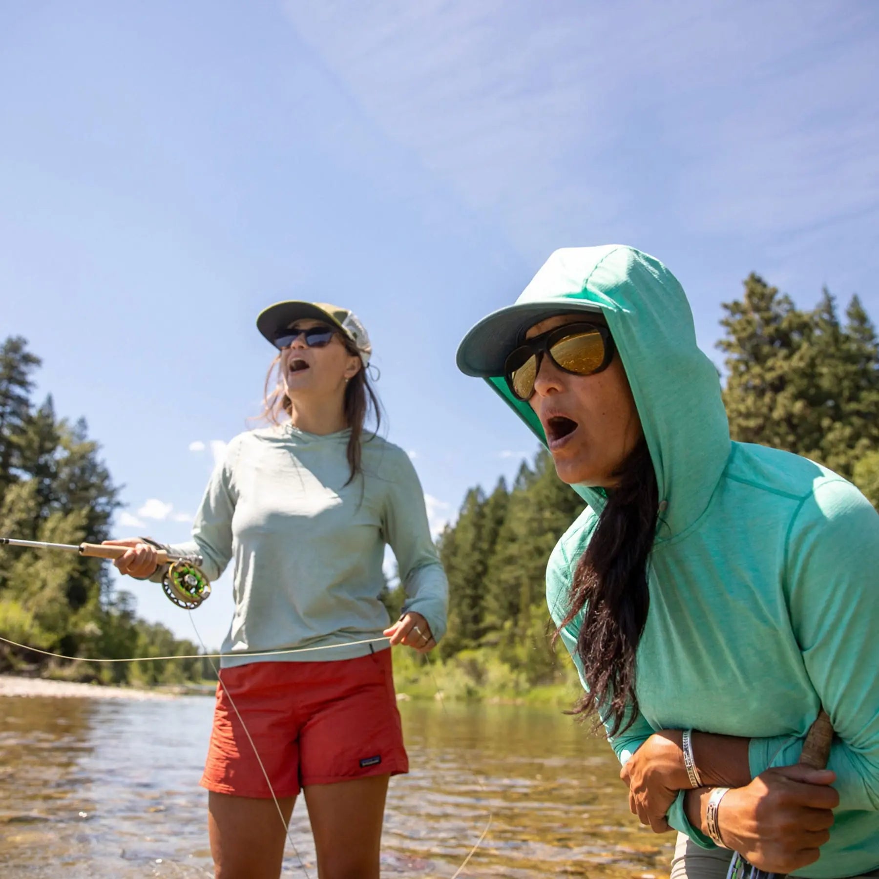 Two women fly fishing by a lake with trees in the background