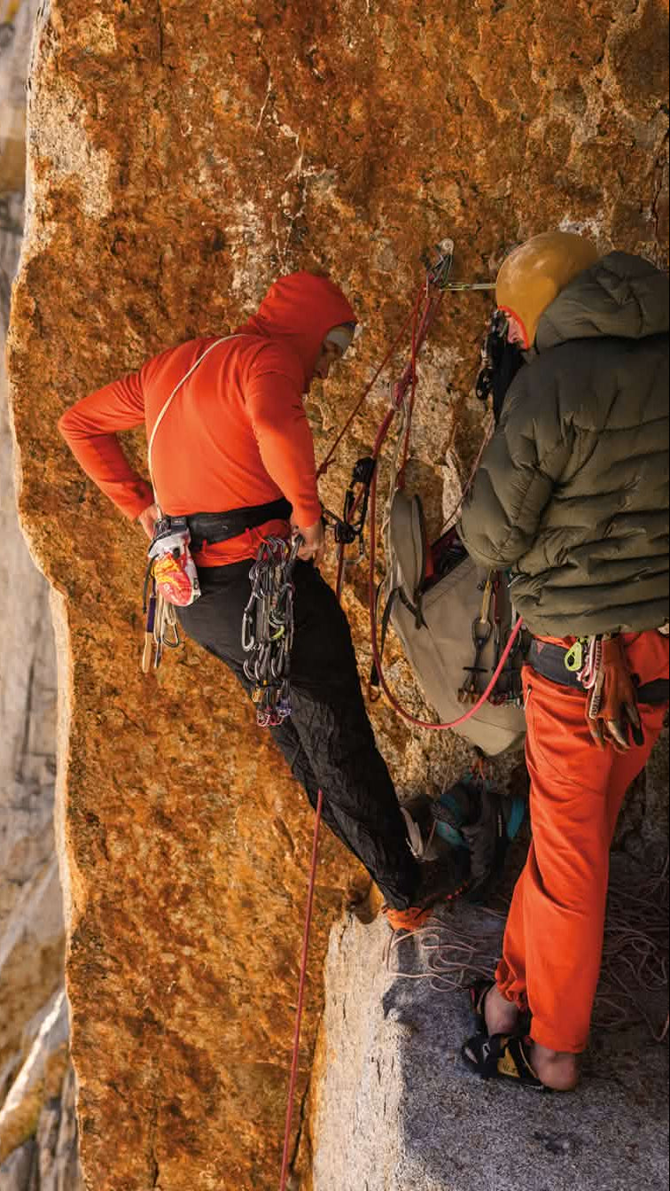 Two climbers on a rocky cliff, one in an orange jacket and the other in a green jacket.