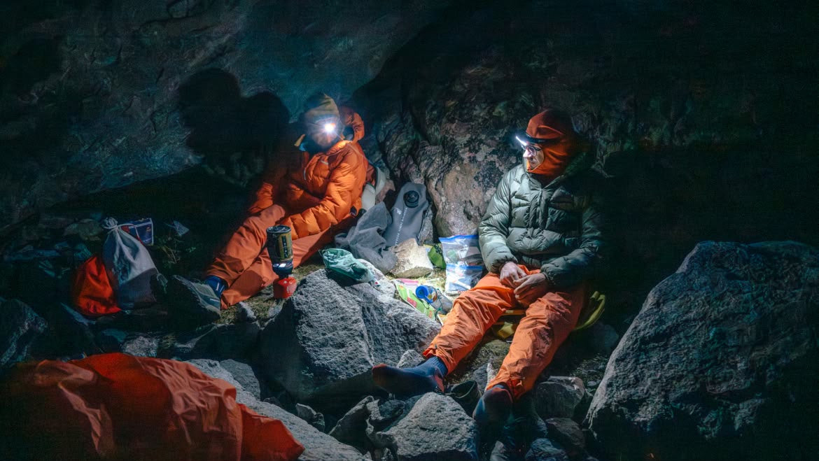 Two people in orange and gray jackets sitting on rocks beside a cliff, illuminated by headlamps.