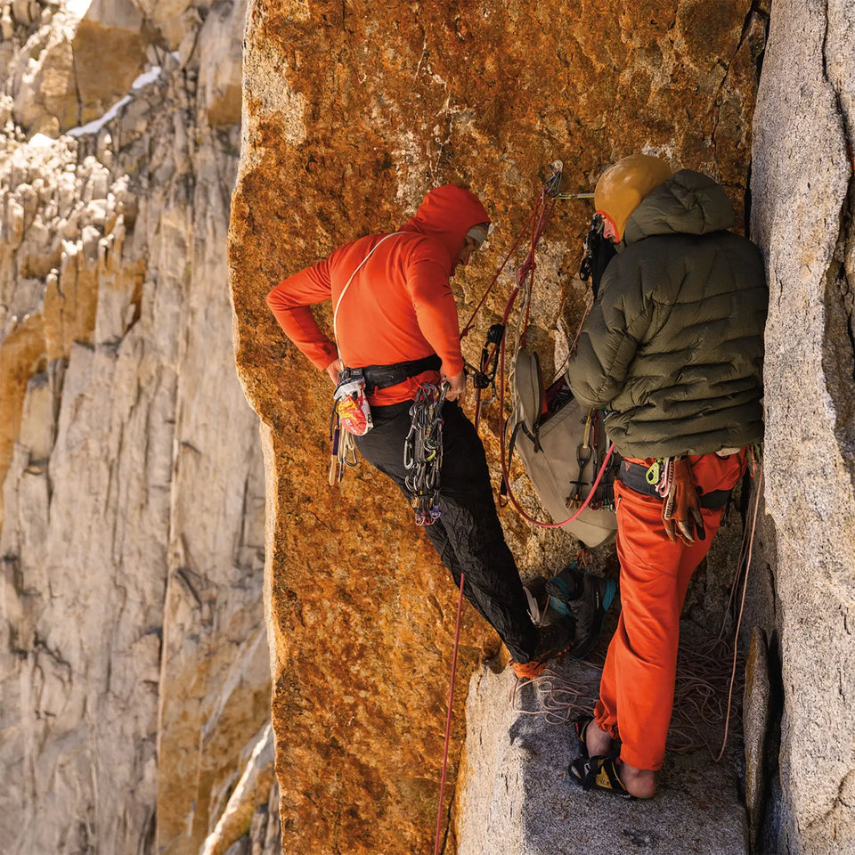 Two climbers on a rocky cliff face, one in orange and the other in green, with climbing gear.
