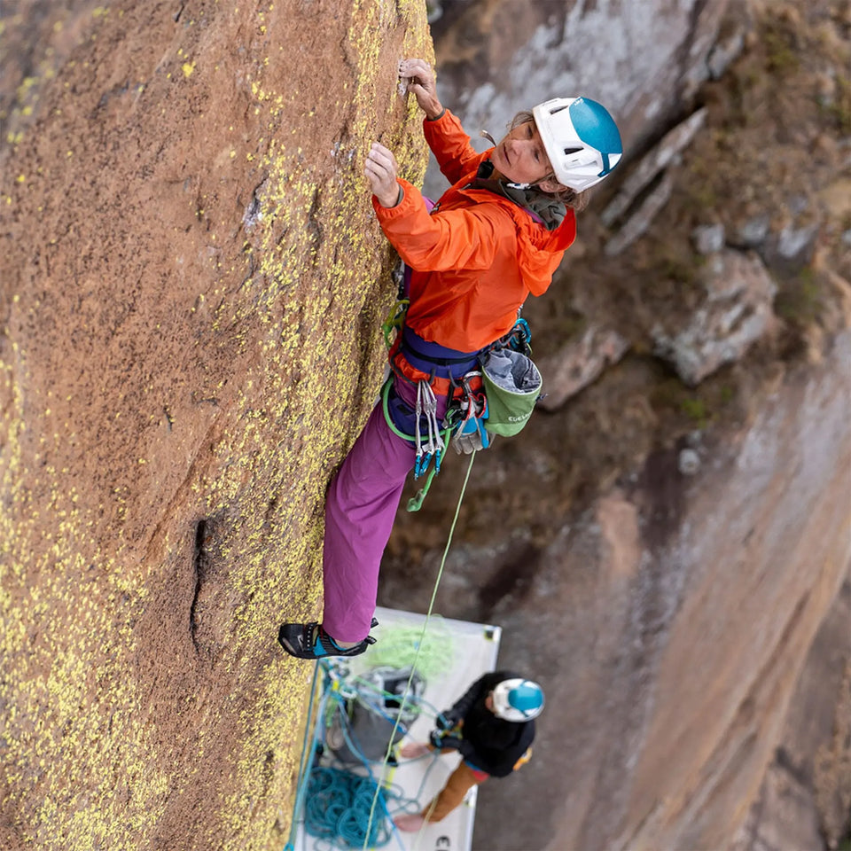 Person climbing a rock wall with another person observing from below.