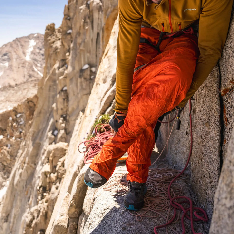 Person in orange climbing pants and yellow jacket on a rocky mountain ledge with climbing gear.