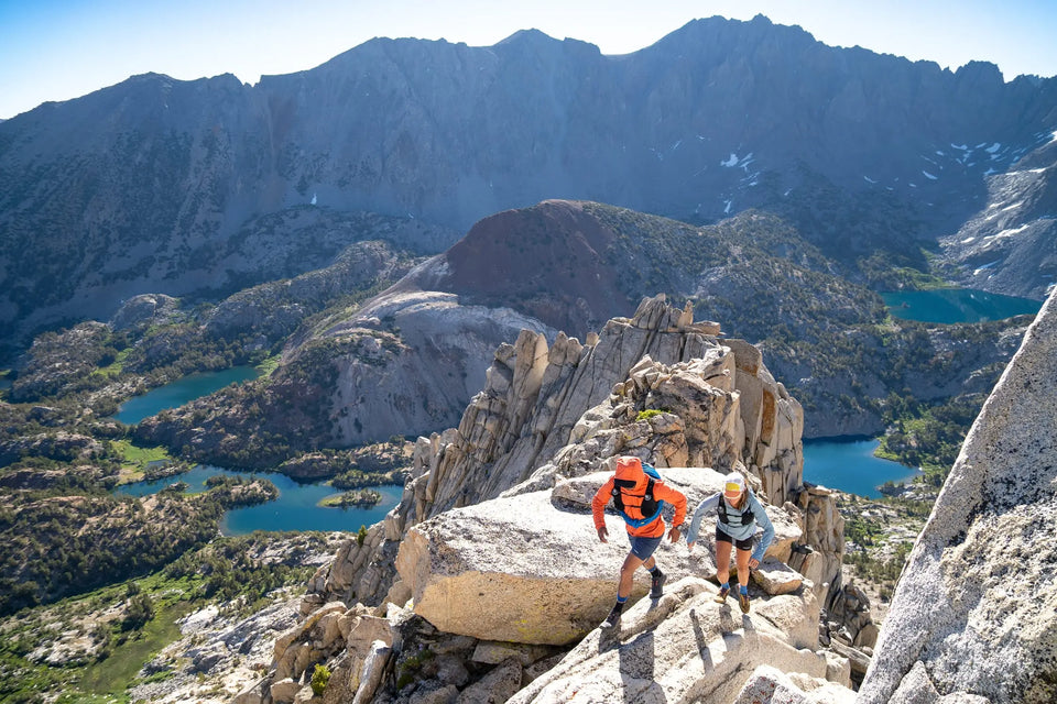 Two hikers on a rocky mountain peak with a scenic view of mountains and lakes below.