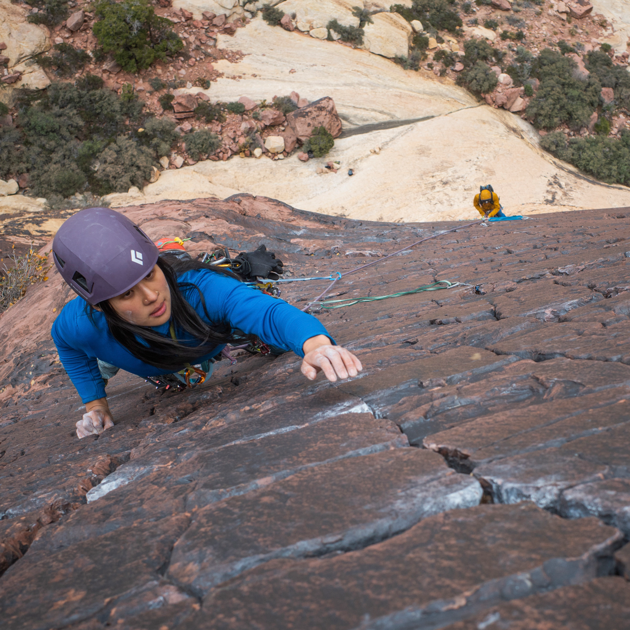 A rock climber wearing a blue jacket, purple helmet, and harness scales a steep reddish-brown rock face in a desert canyon setting.
